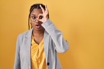 Poster - African american woman with braids standing over yellow background doing ok gesture shocked with surprised face, eye looking through fingers. unbelieving expression.