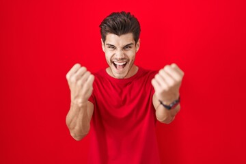 Wall Mural - Young hispanic man standing over red background angry and mad raising fists frustrated and furious while shouting with anger. rage and aggressive concept.