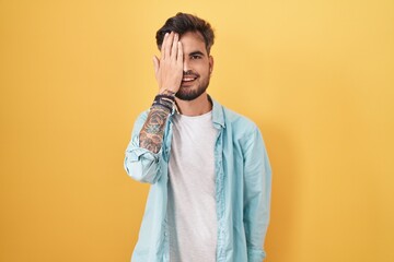 Wall Mural - Young hispanic man with tattoos standing over yellow background covering one eye with hand, confident smile on face and surprise emotion.