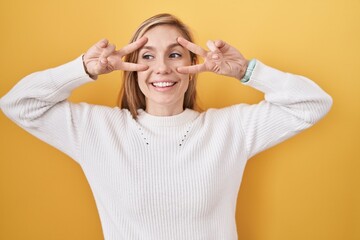 Canvas Print - Young caucasian woman wearing white sweater over yellow background doing peace symbol with fingers over face, smiling cheerful showing victory