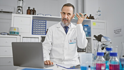 Poster - Hispanic grey-haired middle age man, a lab scientist, engrossed in science research, listening to a voice message on his smartphone at the laboratory table