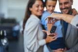 Fototapeta  - Happy parents with small kid after receiving keys for their new car in a showroom. Congratulations, we have a deal about buying a car! Happy family came to an agreement with a car salesperson
