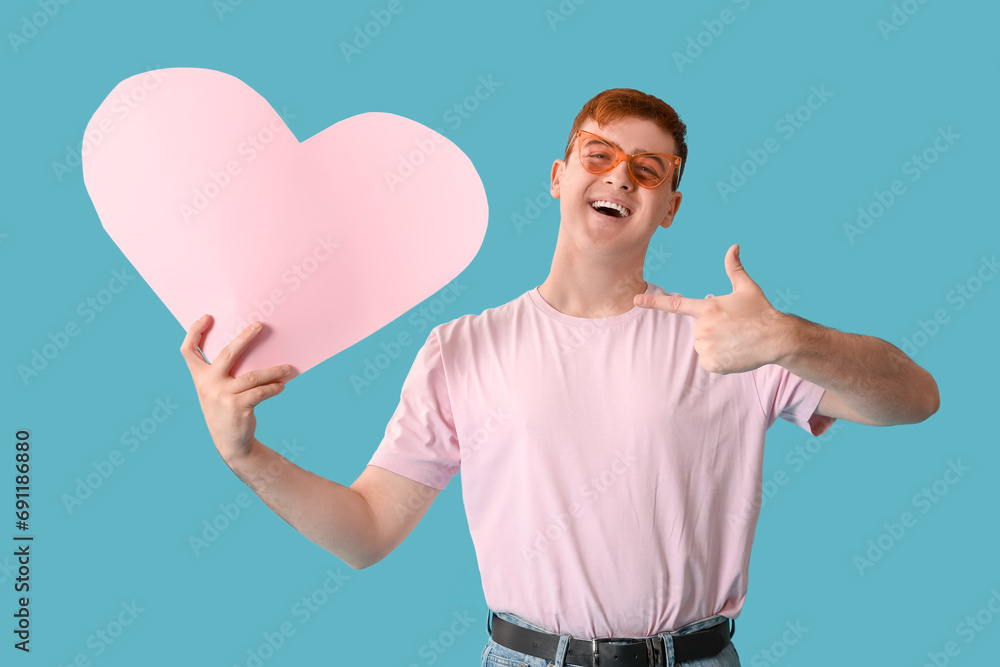 Young man with pink paper heart pointing at something on blue background