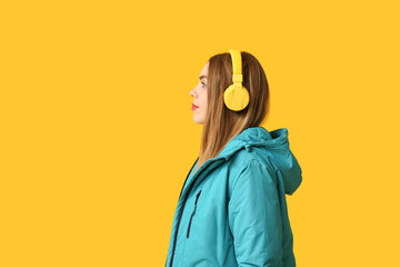 Wall Mural - Beautiful young woman in warm winter clothes with headphones on yellow background