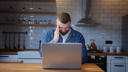 Wall Mural - Bearded young adult man sitting against the kitchen counter having headache while using laptop. Tired young man get having pain from a headache or migraine	