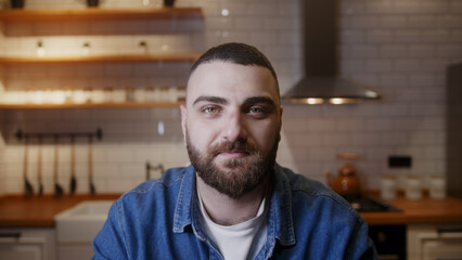 Bearded young adult business man looking at the camera and listening, making conference business call in the kitchen. Video call event by work or study concept	