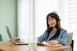 © bongkarn - Young happy and pretty Asian girl wearing headphones is doing homework while studying online at home