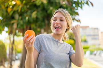 Wall Mural - Young blonde woman holding an orange at outdoors celebrating a victory