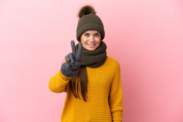 Wall Mural - Young caucasian girl with winter hat isolated on pink background smiling and showing victory sign