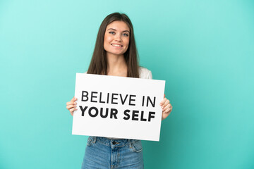 Wall Mural - Young caucasian woman isolated on blue background holding a placard with text Believe In Your Self with happy expression