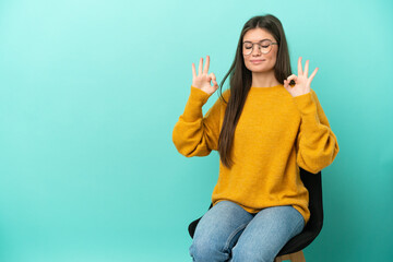 Wall Mural - Young caucasian woman sitting on a chair isolated on blue background in zen pose