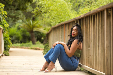 South American woman, young, beautiful, brunette, with colorful top and jeans sitting on a wooden bridge, stressed, overwhelmed and desperate. Concept of beauty, fashion, trend, ethnicity, diversity.