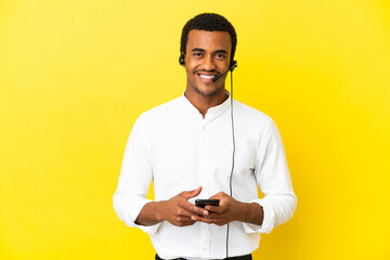 Canvas Print - African American Telemarketer man working with a headset over isolated yellow background sending a message with the mobile