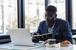 © BUDDHA - Handsome young African man wearing formal suit sitting at a coffee shop with pensive look, thinking of business plans, holding a pen, leaning his elbow on the table, signing papers, working on laptop