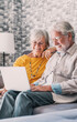 © Daniel - Pretty elderly 70s grey-haired couple resting on couch in living room hold on lap laptop watching movie smiling enjoy free time, older generation and modern wireless technology advanced users concept.