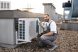 © sofiko14 - Smiling professional worker wearing overalls crouching and holding hand on air conditioner. Experienced man with braided hair working and taking care of device outdoor.