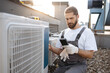 Focused bearded master wearing gloves holding digital multimeter and checking voltage level in air conditioner. Caucasian man working with factory equipment on open air terrace.
