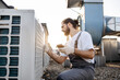 © sofiko14 - Side view of professional builder sitting in front of air conditioner and using measuring device on roof of factory. Caucasian focused male with braided hair testing voltage of climate control unit.