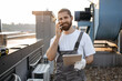 © sofiko14 - Portrait of optimistic adult engineer smiling and talking on modern smartphone while holding tablet in hand. Caucasian male wearing uniform standing on roof of factory and servicing devices.