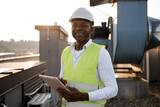 Fototapeta  - Serious factory worker standing and holding modern tablet in hands while servicing devices at factory. African american man wearing uniform inspecting area while performing work in local roof.