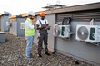 © sofiko14 - African american technician with tablet in hands showing fixed air conditioner to caucasian manager in white hard hat on rooftop. Factory workers cooperating for maintaining modern equipment.
