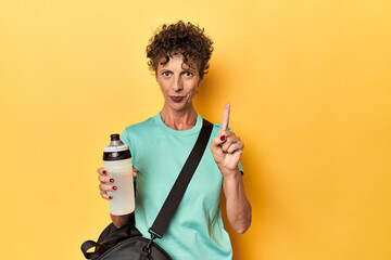 Wall Mural - Sportswoman with gym bag & water on yellow studio showing number one with finger.