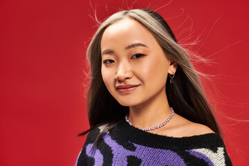 happy and young asian woman with dyed hair in vibrant sweater with animal print posing on red