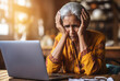 A mature indian woman looks stressed and overwhelmed at a laptop and holds a hand to the head