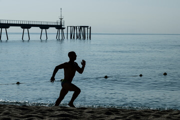 Wall Mural - Running man silhouette training at the beach. Athlete jogging next to the sea with a sea dock in background.