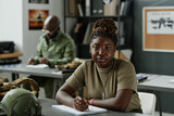 Fototapeta  - African American female student in casualwear sitting by desk with military equipment and looking at camera while making lecture notes