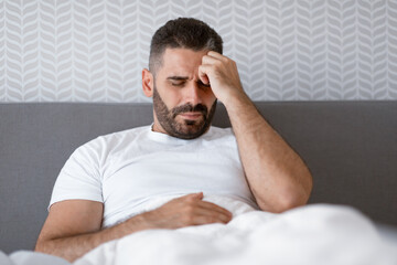 Wall Mural - Unhappy man lying in bed touching head having migraine indoor