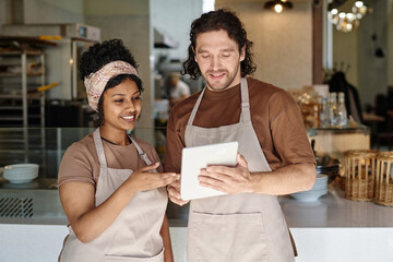 Two biracial bakery workers discussing information on digital tablet screen