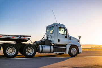 Day cab white local carrier big rig semi truck with step deck semi trailer driving on the road toward the sunset