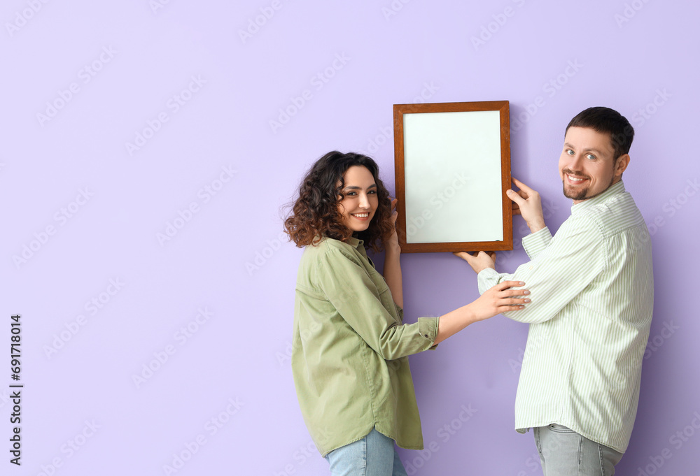 Young couple hanging blank frame on lilac wall