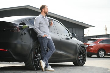 Handsome man drinking coffee while charging electric car