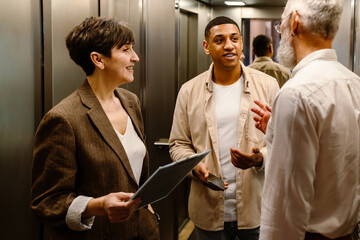 Canvas Print - Three diverse colleagues smiling while discussing business issues in elevator