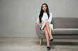 © serhiibobyk - Elegant female doctor resting in comfortable sofa after work day. Low angle view of smiling woman with long dark hair, wearing medical uniform sitting with crossed legs in spacious room. Work concept.