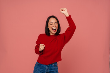 Wall Mural - Excited asian woman screaming with joy and showing winner gesture while standing isolated over pink background