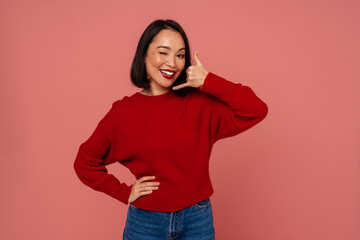 Wall Mural - Cheerful asian woman showing call gesture and winking while standing isolated over pink background