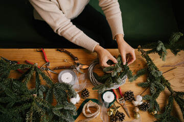 Wall Mural - Woman making Christmas wreath using natural pine branches, sitting near wooden table.