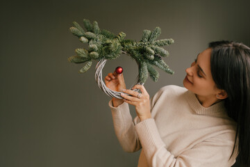 Wall Mural - Woman holding Christmas wreath made of natural pine, standing near grey wall.