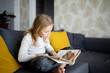 © evgeniia_1010 - On the couch in her house, a little girl with lush blond hair is turning over the pages of a book with interest.