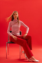 Fashionable confident woman wearing trendy pink turtleneck, classic red trousers, metallic color square toe shoes, sitting on chair, posing on red background. Full-length studio fashion portrait