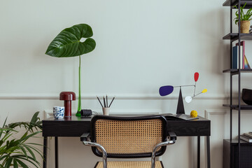 Modern composition of workplace interior with black desk, rattan chair, colorful sculpture, vase with leaves, rack, books, wall with stucco and personal accessories. Home decor. Template.