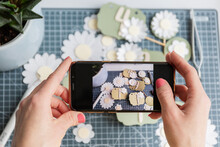 Woman Framing A Creative Display Of Handmade Paper Tags And Daisies Through A Smartphone Screen