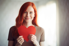 AI Generated Image Of Smiling Redhead Woman With Blue Eyes Holding A Red Paper Heart While Looking At Camera Against Blurred White Background