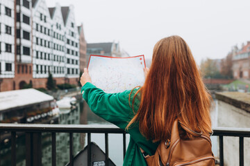 Wall Mural - Attractive young female tourist is exploring city. Redhead girl with backpack holding a paper map on city street in Gdansk. Traveling Europe in autumn, Motlawa river, rear view