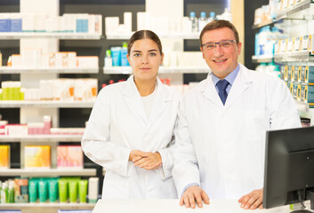 Two smiling female and male pharmacist workers stand shoulder to shoulder and affably welcoming visitors in pharmacy s sales area