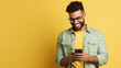 Portrait of a Charming male entrepreneur with hand in pocket smiling and messaging over mobile phone. Happy young man wearing denim shirt and eyeglasses standing isolated on yellow background