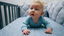 Blonde Baby Boy In Blue Clothes Lies On Belly Leaning On His Hands. Adorable Kid Looks Up With Surprise.